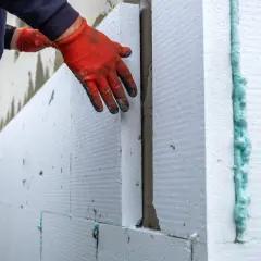 Ręce w czerwonych rękawicach przykładają styropian do ściany. To kluczowy etap prac, by dowiedzieć się, czym ocieplić dom z zewnątrz.