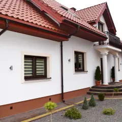 Biały dom z czerwonym dachem i brązowymi rynnami. Na pierwszym planie ozdobny cokół, schody i zadbany ogród.