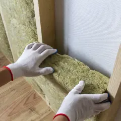 Ręce w białych rękawiczkach dopasowują wełnę mineralną do drewnianej konstrukcji ściany. Do cięcia wełny mineralnej użyj specjalnego noża.