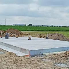 Gotowa płyta fundamentowa na budowie, otoczona piaskiem i zielenią. W tle widać budynki przemysłowe.