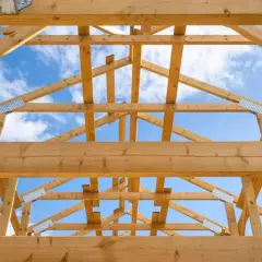 Drewniana więźba wieszarowa tworzy konstrukcję dachu, widoczną na tle błękitnego nieba z chmurami.