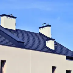 Nowoczesny dach bez okapu z ciemnogranatową dachówką i białymi kominami na tle błękitnego nieba.
