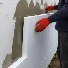 Ręce w czerwonych rękawiczkach przytwierdzają płyty styropianowe do ściany. Trwa docieplenie elewacji.