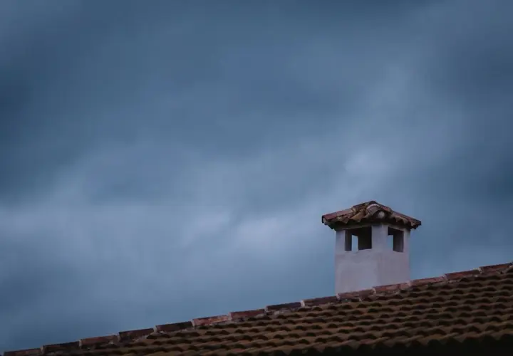 Komin na dachu, jakby pokryty folią w płynie, na tle burzowego nieba.