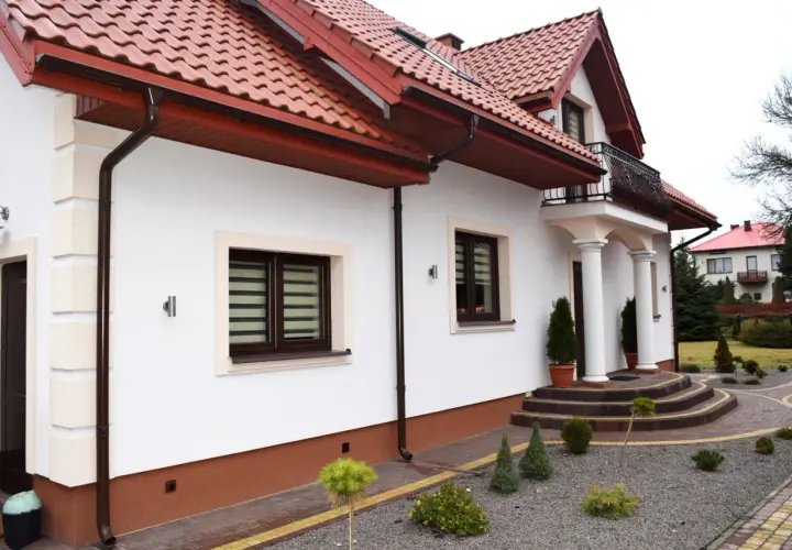 Biały dom z czerwonym dachem i brązowymi rynnami. Na pierwszym planie ozdobny cokół, schody i zadbany ogród.