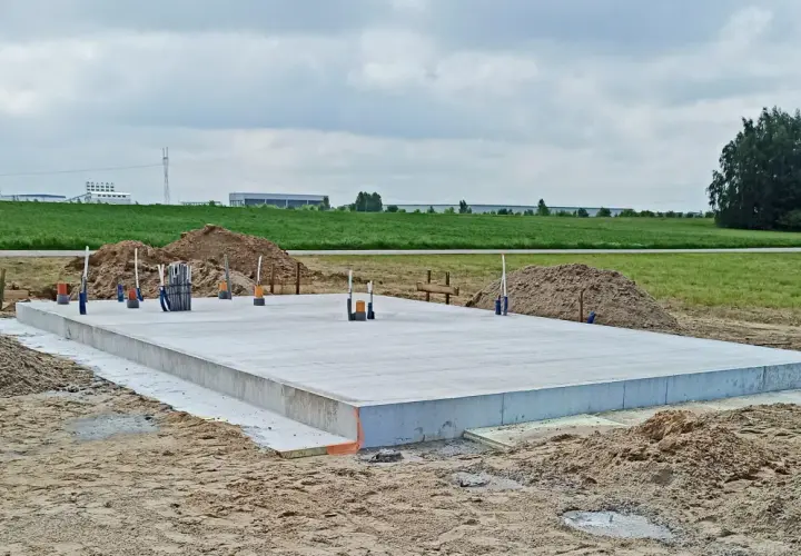 Gotowa płyta fundamentowa na budowie, otoczona piaskiem i zielenią. W tle widać budynki przemysłowe.