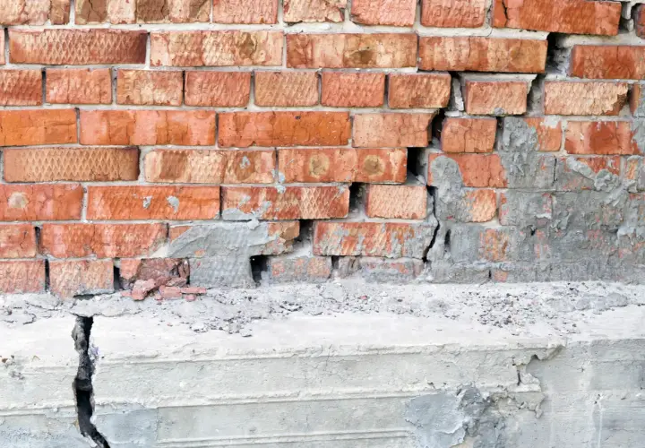 Uszkodzona ceglana ściana z widocznym pęknięciem w betonie, sugerującym podcinanie fundamentów.