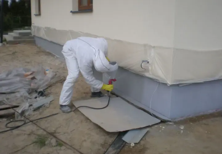 Pracownik w kombinezonie wykonuje malowanie elewacji po ociepleniu styropianem, zabezpieczając dolną część budynku folią.