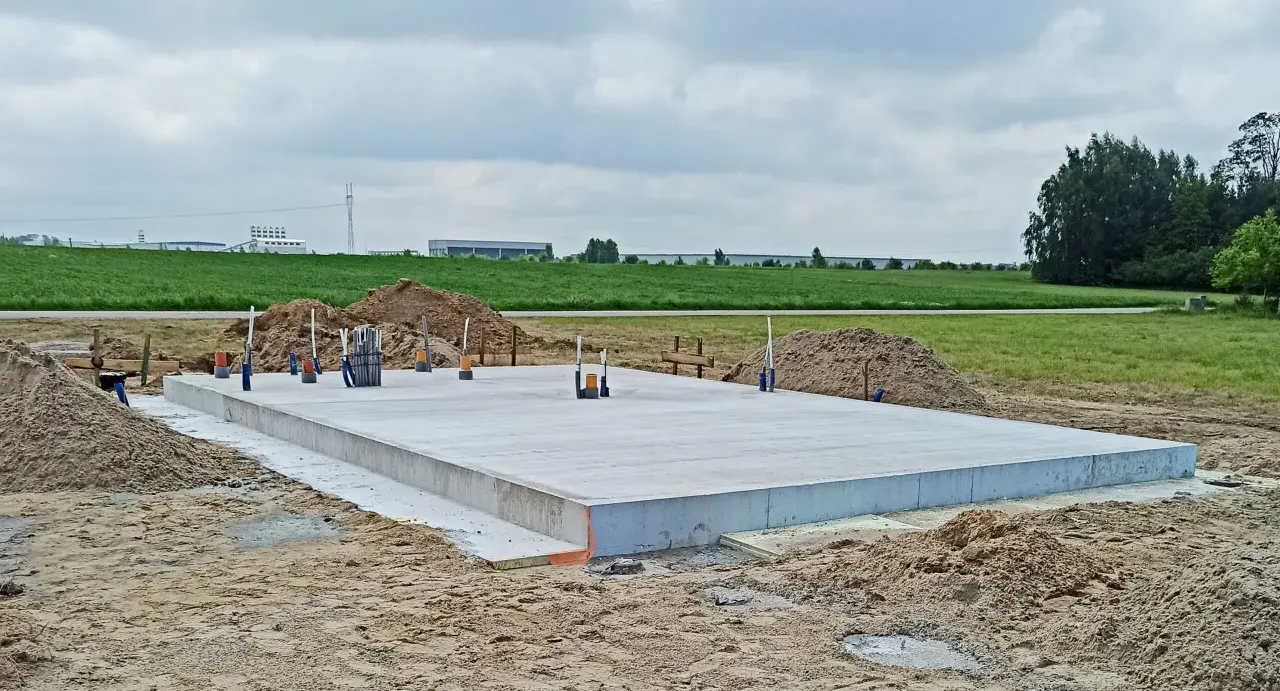 Gotowa płyta fundamentowa na budowie, otoczona piaskiem i zielenią. W tle widać budynki przemysłowe.