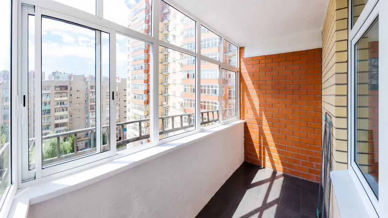 Jasny balkon typu loggia z dużymi oknami, widokiem na budynki mieszkalne i ceglaną ścianą.
